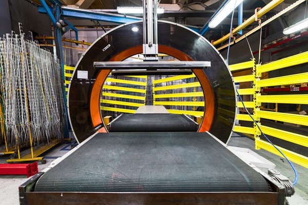 Orbital wrapping machine with a conveyor belt and surrounding yellow bars in a factory setting.