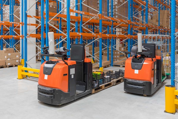 The image shows two orange electric tow tugs in a warehouse, positioned in front of blue and orange shelving racks filled with boxes.