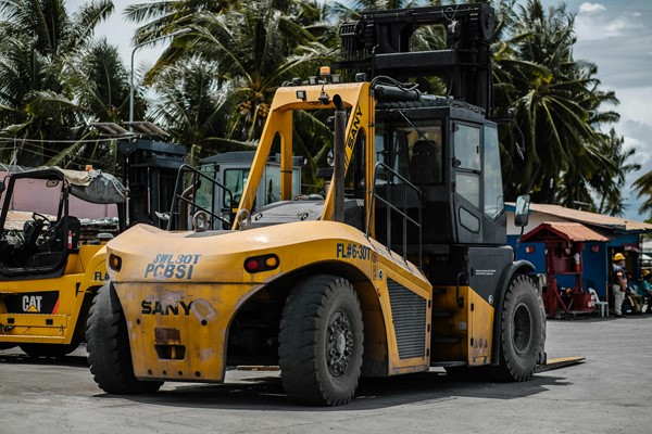 A large yellow forklift is parked outdoors, with palm trees and other equipment in the background.