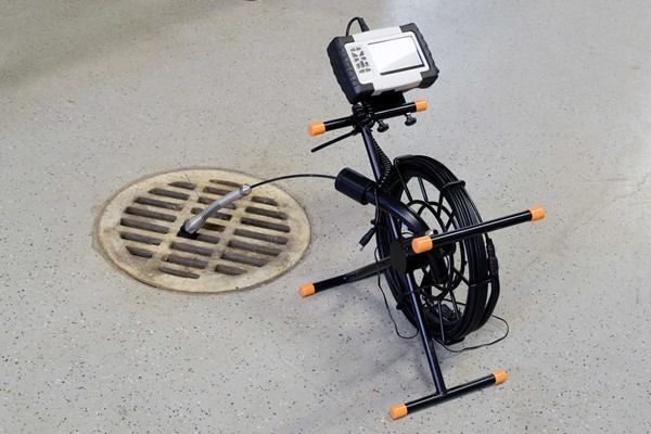 A sewer inspection camera inserted into a drain, with a connected monitor and cable reel on the floor.