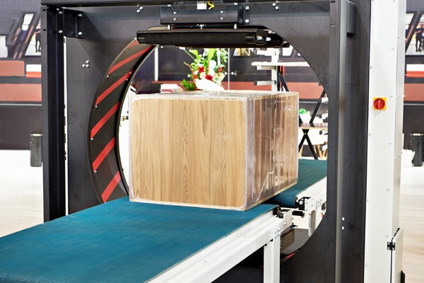 A large wooden box wrapped in plastic film is being processed on a blue conveyor belt under an orbital wrapping machine.