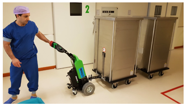 A person in blue scrubs uses a green electric tug to move metal carts in a hospital hallway.