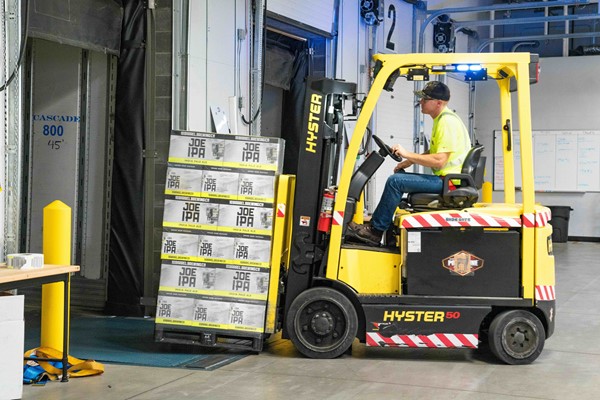 A worker in a safety vest and cap drives a yellow forklift, moving a pallet of boxed beer into a loading dock inside a warehouse.