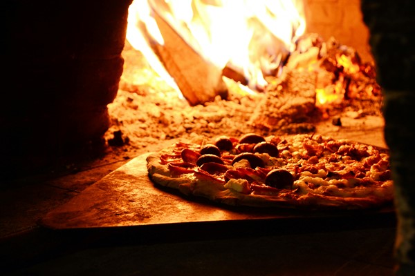 A pizza baking in a wood-fired oven with flames and embers glowing in the background.