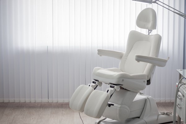 A modern white podiatry chair with adjustable footrests and armrests, positioned in a bright room with vertical blinds.