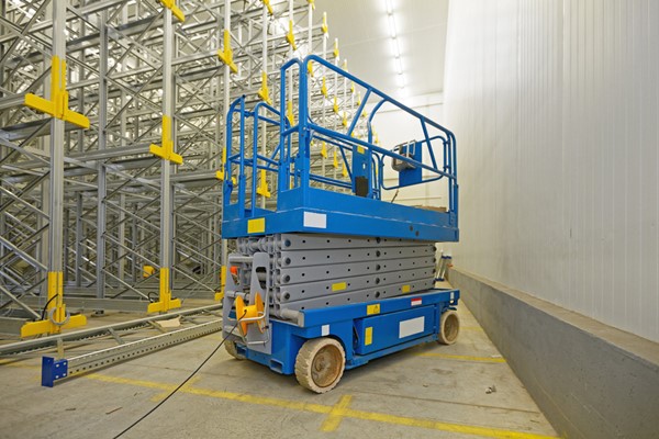 A blue scissor lift in an industrial warehouse, surrounded by metal racking and yellow safety markers.