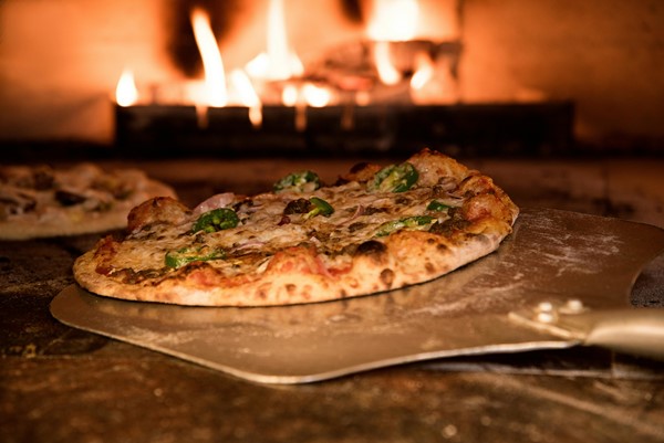 A wood-fired oven with a blazing fire, baking a pizza topped with jalapeños and cheese. The pizza is being pulled out with a metal peel, showing a crispy, charred crust.