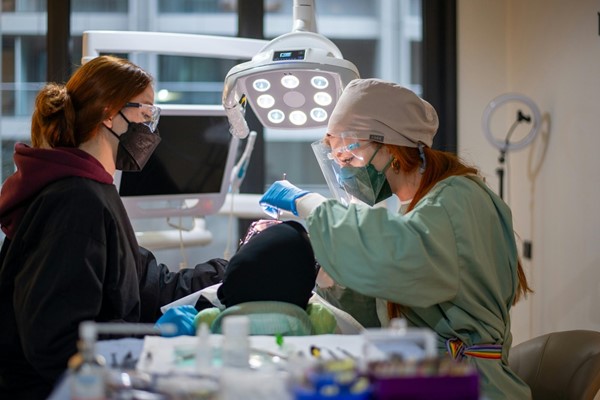 Two dental professionals wearing masks and protective gear working on a patient under a bright overhead light.