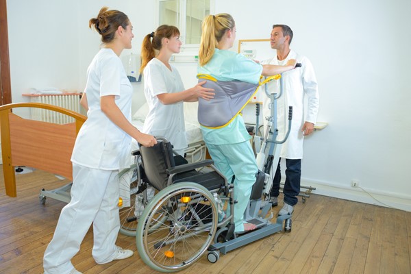 Healthcare professionals assist a patient using a standing aid lift from a wheelchair in a medical setting.