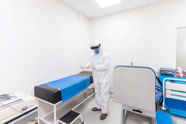 A healthcare professional in full protective gear prepares for a procedure in a clean, well-equipped examination room with a medical bed.