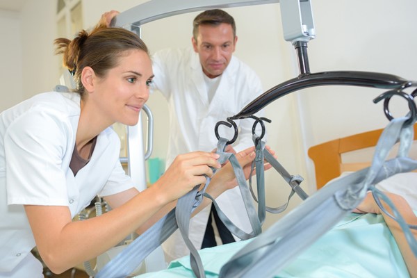 Two healthcare professionals adjust the straps of a patient lift system in preparation to assist a patient in a medical facility.