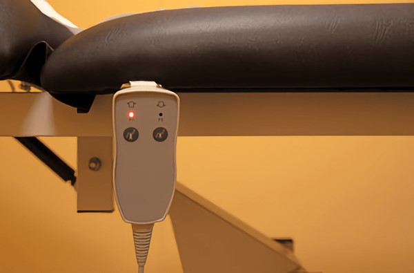 Close-up of an electric treatment table's control panel with a red light, black cushion, and metal frame against a yellow background.