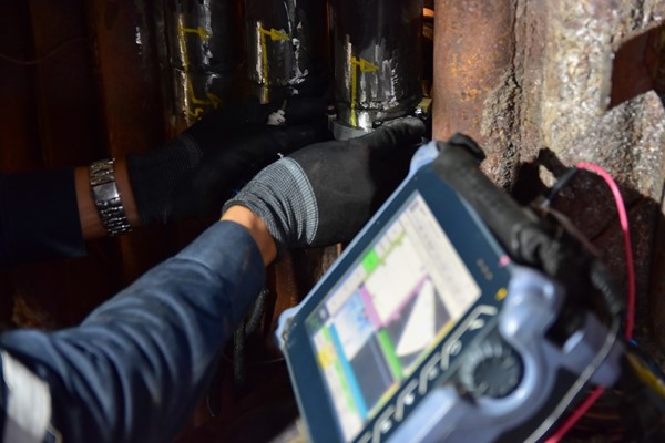 A technician wearing protective gloves uses an inspection device to examine industrial pipes. The device's screen displays data, and the setting appears to be a worn, industrial environment.